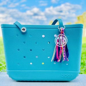 May include: A turquoise blue tote bag with a white monogram charm and a colorful tassel. The bag has a  "BOGG" logo on the bottom.