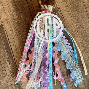 May include: A white acrylic keychain with a circle charm featuring the monogram "GAP" in white lettering. The charm is attached to a colorful tassel made of various ribbons and trims in shades of pink, blue, yellow, and purple.