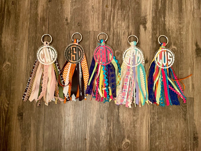May include: Five colorful keychains with different initials on each. The keychains are made of clear acrylic with a white circle around the initials. The keychains are attached to a silver ring and have a variety of colorful ribbons attached to the ring.