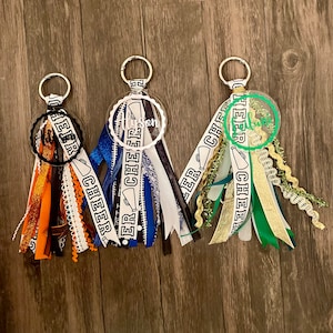 May include: Three white and black cheer keychain tassels with clear round charms. The charms have the word "cheer" written on them. The tassels are made of various ribbons and have a gold, blue, and green color scheme.