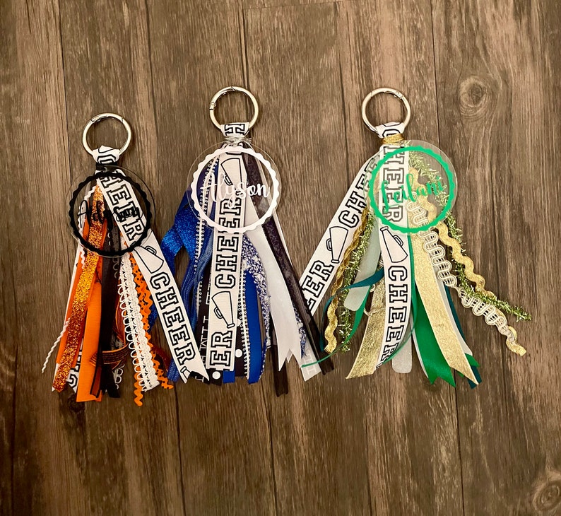 May include: Three white and black cheer keychain tassels with clear round charms. The charms have the word "cheer" written on them. The tassels are made of various ribbons and have a gold, blue, and green color scheme.