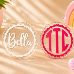 May include: Two circular clear acrylic ornaments with scalloped edges. The left ornament features the white text Bella in a script font. The right ornament displays the hot pink monogram letters TTC in a bold, block-style serif font.