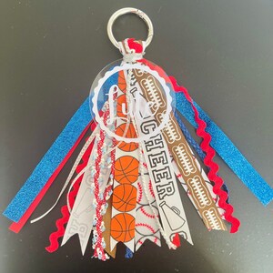 May include: A white acrylic keychain with the word "Little Cheer" written in black. The keychain has a variety of ribbons attached, including blue, red, white, brown, and a white ribbon with basketball and baseball designs.