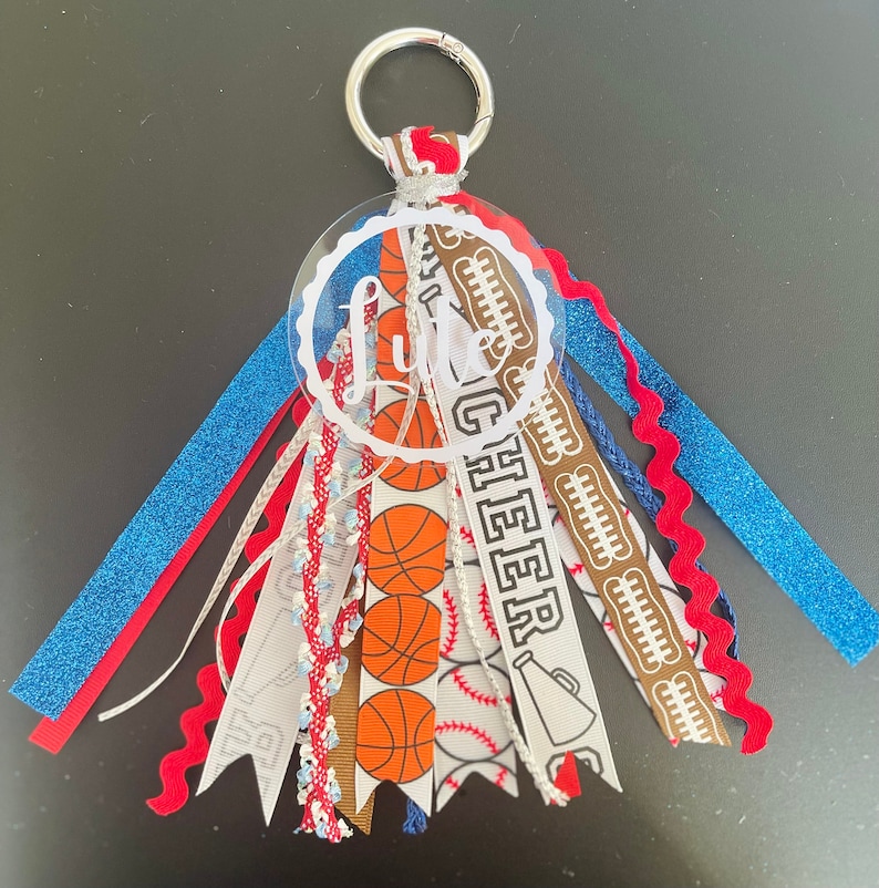 May include: A white acrylic keychain with the word "Little Cheer" written in black. The keychain has a variety of ribbons attached, including blue, red, white, brown, and a white ribbon with basketball and baseball designs.