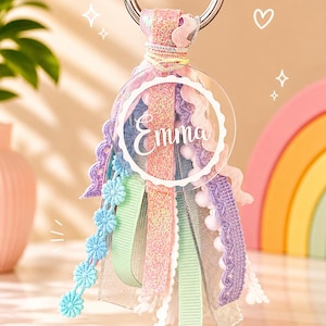 Pode incluir: Um porta-chaves com um pendente circular de acrílico transparente com o nome Emma impresso em branco. O pendente está preso a um anel de metal prateado e decorado com uma borla texturizada feita de fitas em tons pastel rosa, azul, roxo e verde menta.