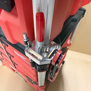 May include: A red and black Milwaukee Packout tool box with a silver metal accessory. The accessory holds a red Sharpie marker, a black pen, and a red tool. The image is taken from a close-up angle.