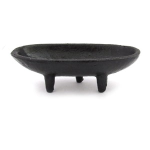 May include: A black cast iron oval dish with three short legs. The dish is empty and has a smooth, slightly textured surface.