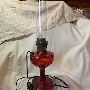 May include: A red glass oil lamp with a clear glass chimney and a black electrical cord. The lamp is sitting on a clear glass plate.