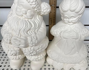 May include: White ceramic Santa Claus and Mrs. Claus figurines. Santa is holding a bag and Mrs. Claus is holding a stocking. Both figurines have a detailed design.