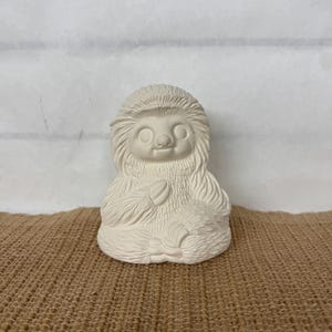 May include: White ceramic figurine of a sloth sitting in a meditative pose with its arms crossed in front of it. The sloth has a detailed face with large eyes and a small mouth.
