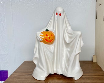 Puede incluir: Una figurita de fantasma de cerámica blanca que sostiene una pequeña calabaza naranja con una cara tallada. El fantasma tiene ojos rojos y lleva una sábana blanca.