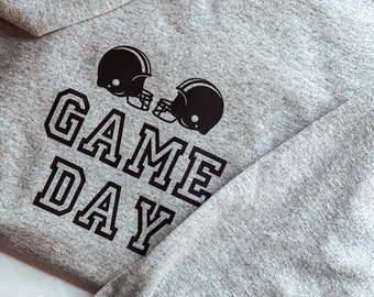 Puede incluir: Sudadera gris con texto negro que dice "GAME DAY" y dos cascos de fútbol negro encima del texto.