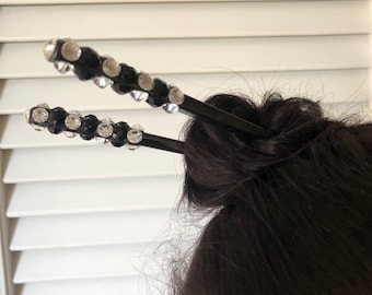 May include: A pair of black hair sticks with clear rhinestone accents. The sticks are crossed and used to hold a bun of hair.