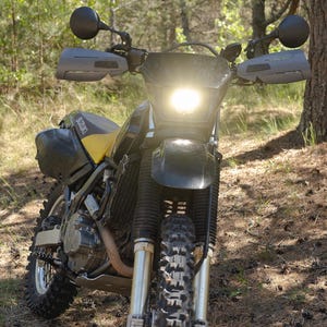 Może przedstawiać: Czarno-żółty motocykl typu dual-sport marki Suzuki z jasnym reflektorem, oponami terenowymi, osłonami dłoni i bocznymi torbami. Motocykl posiada wysoki przedni błotnik, metalowe przednie widelce oraz czarne siedzenie z logo Suzuki.