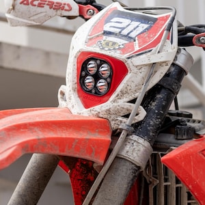 Op de afbeelding: Close-up van een rode en witte crossmotor, bedekt met modder. De motor heeft een opvallende voorspatbord, een koplamp met vier lampen en het nummer 211. Het stuur toont "Acerbis" branding.