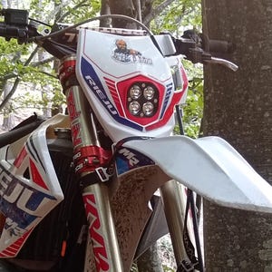 May include: A Rieju dirt bike with a white, red, and blue color scheme is parked in a forest. The bike has a white front fender, a headlight with four lights, and the Rieju logo is visible on the front and side.