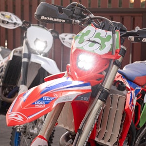 May include: A red and white dirt bike with a bright headlight and the number 353 on the front. The bike has black tires and a blue seat. The image also shows a second dirt bike in the background.