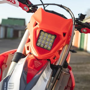 May include: Close-up of a red and white motorcycle with a bright LED headlight. The front fairing features the text "MST" and "www.mstrally.com". The handlebars have "Acerbis" branding.
