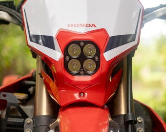 May include: Close-up of a red and white Honda motorcycle headlight assembly. The headlight features four round lights and the Honda logo. The assembly is mounted on a red motorcycle frame with gold-colored suspension forks.