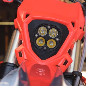 May include: Close-up of a motorcycle headlight assembly. The assembly is primarily bright orange with a black center housing four yellow LED lights. The motorcycle's front suspension and handlebars are also visible.