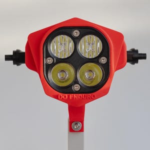 Op de afbeelding: Een rode LED-lichtassemblage met vier heldere lichten. Het licht is gemonteerd op een grijze standaard met een kruisvormige basis. De tekst "DO ENDURO" is gedrukt op de rode behuizing. De lichtassemblage heeft twee zwarte handvatten aan weerszijden.