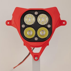 May include: A red LED headlight assembly with four lights. The assembly is mounted on a gray stand. The headlight has two pairs of lights, with the top pair having a different design than the bottom pair.