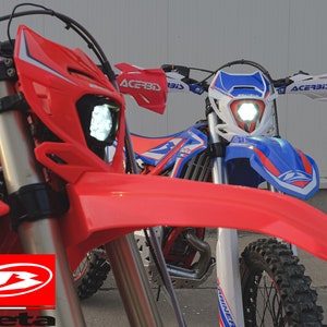 May include: Two off-road motorcycles with bright LED headlights. One is primarily red, the other blue and white. The red motorcycle has the Beta Motorcycles logo. Both bikes have knobbly tyres and handguards.
