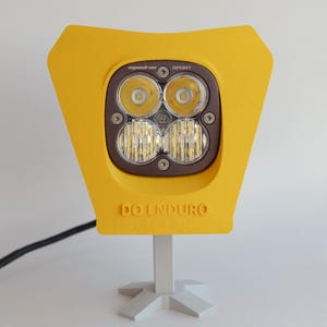 May include: A yellow motorcycle headlight with the text "DO ENDURO" is mounted on a white stand. The headlight has a dark brown center with four circular lights and a series of smaller lights below. A black wire extends from the back.