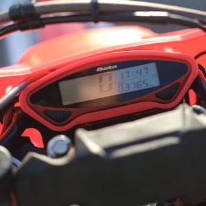 May include: Close-up of a motorcycle dashboard with a red casing. The digital display shows the time, 17:47, and the mileage, 03765. The brand name "Beta" is visible on the dashboard.