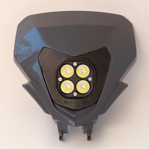 May include: A dark grey motorcycle headlight assembly featuring four bright LED lights arranged in a square. The headlight has a sleek, angular design. The text "DO ENDURO" is visible on the lower section.
