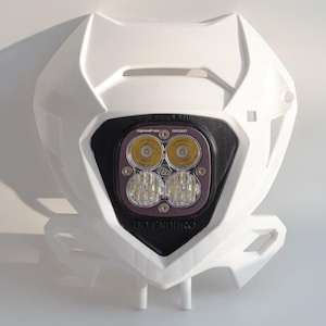 Op de afbeelding: Witte motorfiets koplamp montage met een zwarte en paarse lichtbehuizing. De koplamp heeft vier LED-lampen en de tekst "DO ENDURO". De montage is ontworpen voor offroad-motorfietsen.