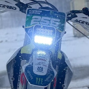 Peut inclure: Gros plan d'une moto tout-terrain Husqvarna avec le numéro 255 bien en évidence. La moto est équipée d'un phare LED lumineux et est recouverte de neige. Le texte "VLADIMIRS ELSTERS" est visible.