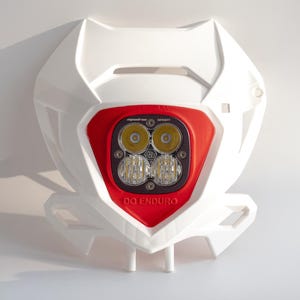 Op de afbeelding: Witte en rode motorfiets koplamp montage met twee paar ronde lichten en de tekst "DO ENDURO". De montage heeft een witte kunststof behuizing en een rood middengedeelte.