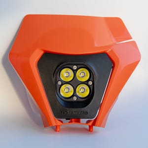 May include: An orange motorcycle headlight assembly with four bright LED lights. The black center section has the text "DO ENDURO" printed on it. The headlight is designed with a geometric shape.
