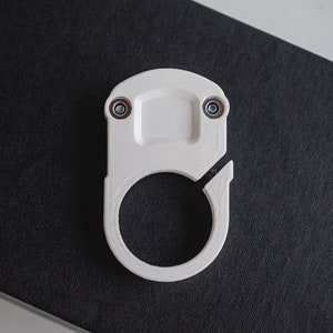 May include: A white, 3D-printed object with a circular opening and a rectangular top, secured with two small screws. The object is set against a dark, textured background, suggesting a modern design.