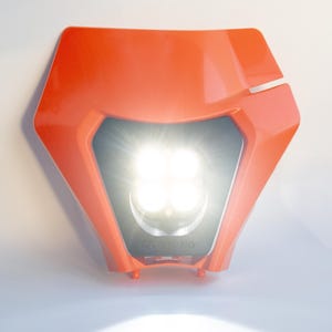 May include: Bright orange motorcycle headlight assembly with four bright LED lights. The headlight has a black center section and a distinctive angular design. The light is turned on, emitting a strong, focused beam.