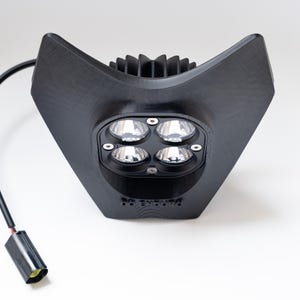 Peut inclure: Phare LED noir avec quatre lumières vives. Le boîtier est de conception angulaire noir mat avec un dissipateur thermique sur le dessus. Un fil noir et un connecteur s'étendent du bas. Le texte "RR ENDURO" est visible.