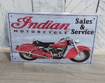 May include: A rectangular metal sign featuring a red Indian motorcycle illustration on a light grey wood-grain background. The text reads Indian Motorcycle Sales & Service in black and red lettering, with small holes in the corners for mounting.