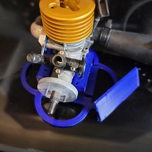 Puede incluir: Un motor modelo de oro y plata con un soporte de plástico azul. El motor tiene una culata dorada y un cigüeñal plateado. El soporte tiene una base circular y tres patas.