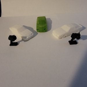 May include: Three miniature toy cars, one green and two white, with black stands. The cars are all facing forward and the stands are positioned behind the cars.