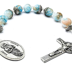May include: A silver rosary with blue and white marbled beads. The rosary has a silver crucifix with a figure of Jesus Christ and a silver medal with the image of Saint Cecilia and the text "SAINT CECILIA".