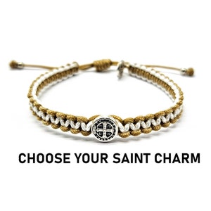 Peut inclure: Un bracelet tressé marron et blanc avec un charme argenté en forme de croix. Le bracelet est réglable et possède une fermeture à nœud coulissant. "Choisissez votre charme de saint" est écrit sous le bracelet.