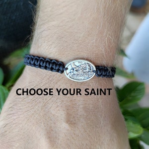 May include: A black braided bracelet with a silver oval charm featuring a religious image. The charm has the words "Italy" and "Ave Maria" inscribed on it. The text "Choose Your Saint" is visible below the bracelet.