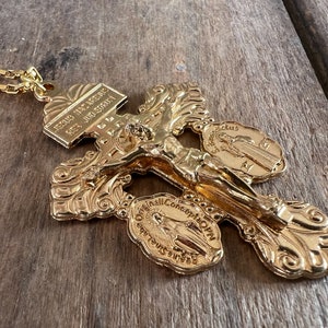 Puede incluir: Un colgante de metal dorado en forma de crucifijo con una cadena. El crucifijo presenta una figura de Jesucristo en la cruz, con tres medallones que representan a la Virgen María. Los medallones tienen inscripciones en latín. El colgante está sobre una superficie de madera.