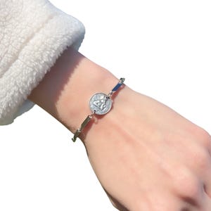 Op de afbeelding: Zilverkleurige armband met een ronde bedel met een engelontwerp. De armband heeft een fijne ketting met rechthoekige schakels. De bedel en schakels zijn zilver van kleur. De armband wordt om een pols gedragen.