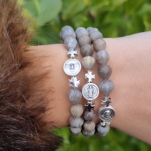 Puede incluir: Tres pulseras de cuentas con cuentas de madera grises y marrones y dijes religiosos plateados. Los dijes incluyen cruces y medallones con figuras religiosas. Las pulseras se usan en una muñeca con una manga de piel marrón.