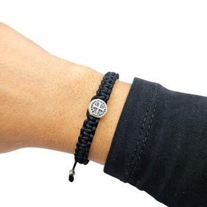 Puede incluir: Un brazalete trenzado negro con un dije de plata con una medalla de San Benito. El dije tiene una cruz y las palabras "EGO VOX" e "IN HOC SIGNO VINCES".