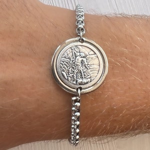 Op de afbeelding: Een zilveren armband met een ronde hanger met een gedetailleerd beeld van een persoon die een berg beklimt. De hanger is bevestigd aan een zilveren ketting.