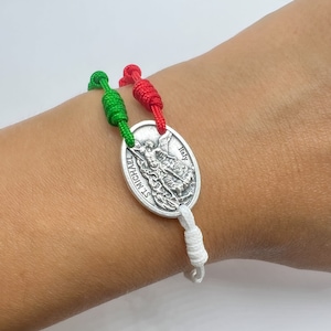 Puede incluir: Un brazalete trenzado blanco, rojo y verde con un dije ovalado de plata que presenta una imagen de San Miguel Arcángel. El dije está inscrito con "St. Michael" e "Italia".