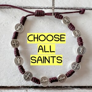 May include: A brown cord bracelet with silver medallions featuring religious imagery. The medallions have a cross and a figure of a saint. The bracelet has the text "CHOOSE ALL SAINTS" printed on a yellow background.